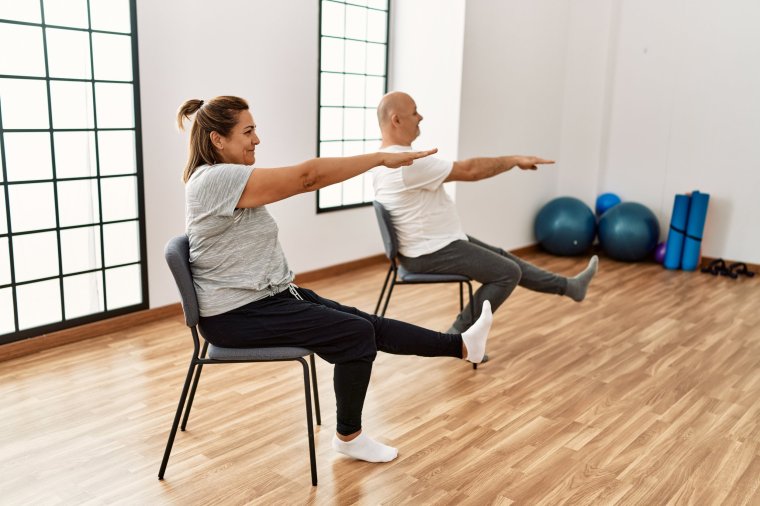 A middle-aged Hispanic couple stretches with a chair in a sports center.